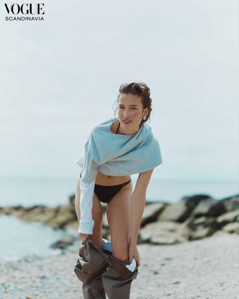 A young woman is on a beach, removing dark olive-green waders. She wears a pale turquoise cropped top, a black bikini bottom, and has wet hair.