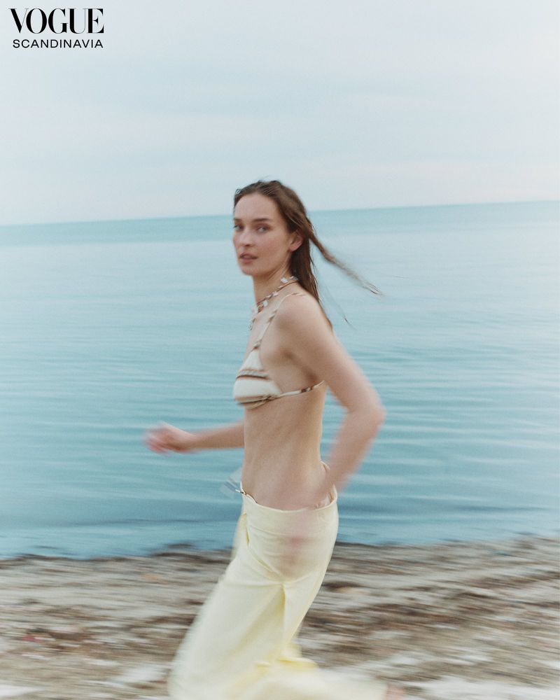 Woman in a bikini top and yellow pants walks on a sandy beach with a blurred motion effect, Vogue Scandinavia logo in the upper left.