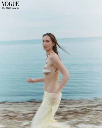 Woman in a bikini top and yellow pants walks on a sandy beach with a blurred motion effect, Vogue Scandinavia logo in the upper left.