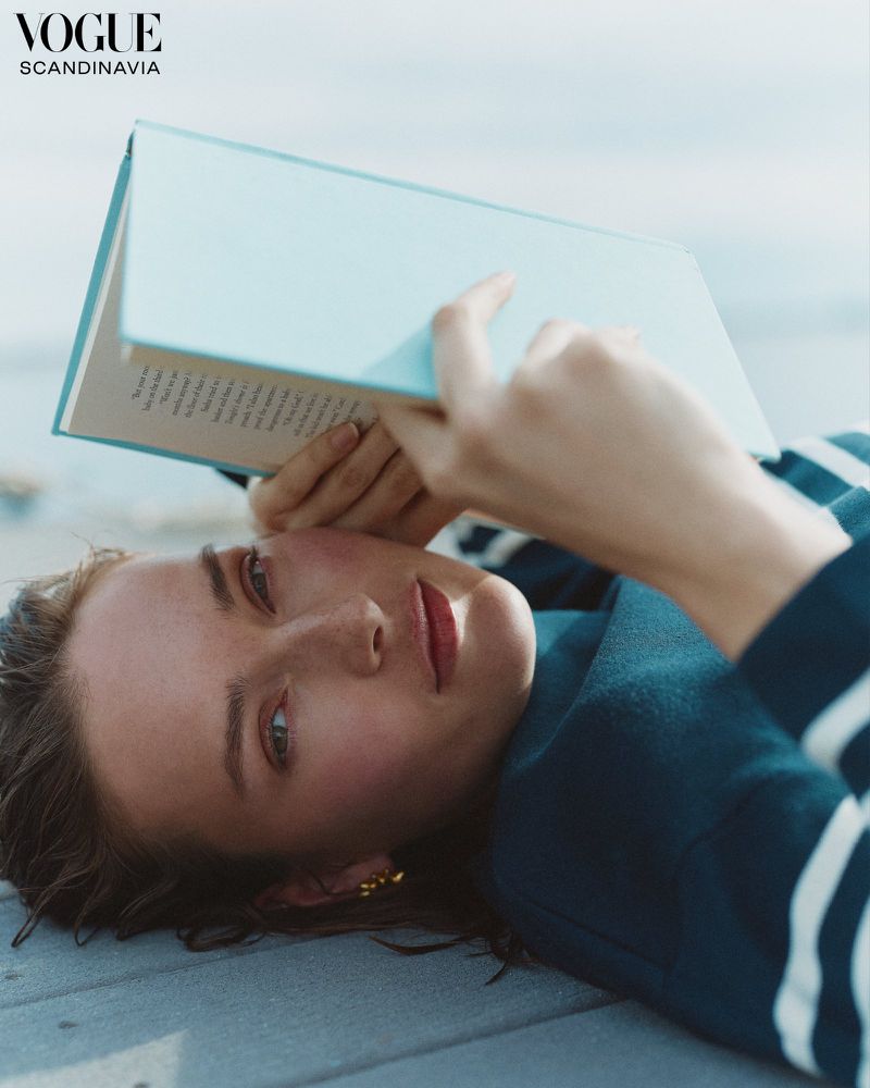 Woman reading a book while lying down outside on wooden planks; she is wearing a navy blue and white striped sweater, and golden earrings.