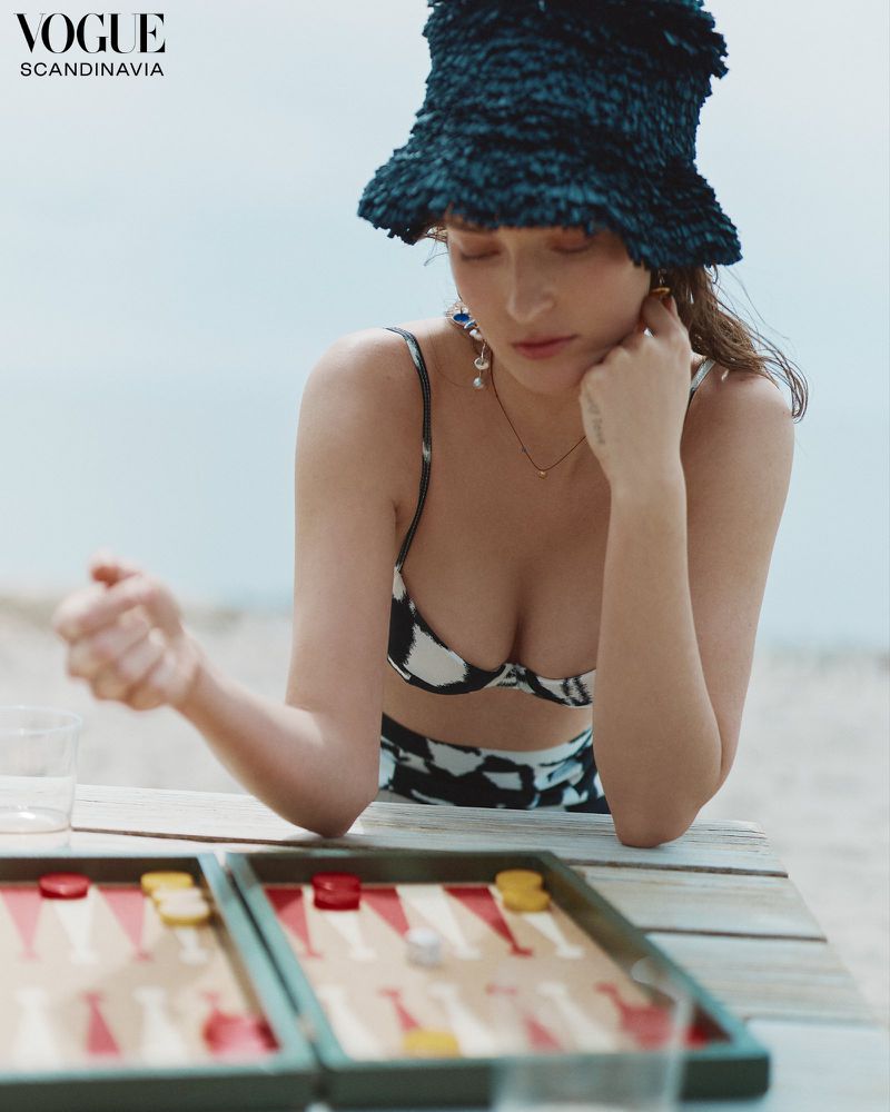 A woman wearing a printed bikini and fuzzy blue bucket hat plays a game of backgammon on a weathered wooden table against an outdoor beach backdrop.