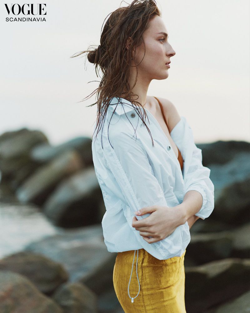 Woman in white shirt and gold skirt on rocky coast with wet hair in editorial fashion shot for Vogue Scandinavia, looking contemplative.