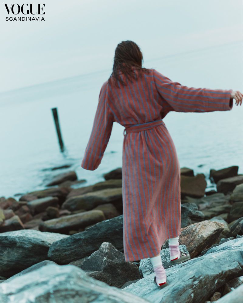 An adult female balances on rocks by the ocean in a striped pink robe. She faces away from the camera and towards the sea.