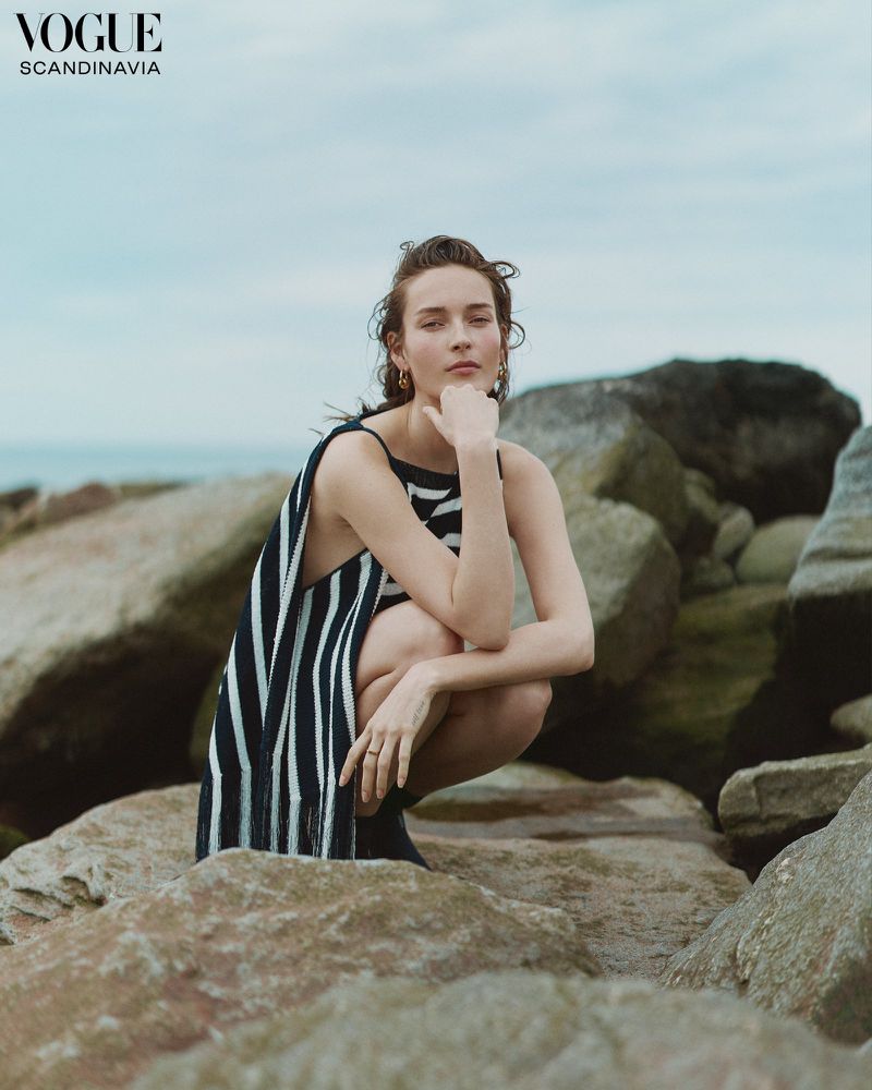 A woman in a striped dress squats on rocks near the ocean, looking at the camera, with a cloudy sky in the background. Vogue editorial.