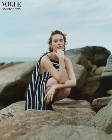 A woman in a striped dress squats on rocks near the ocean, looking at the camera, with a cloudy sky in the background. Vogue editorial.