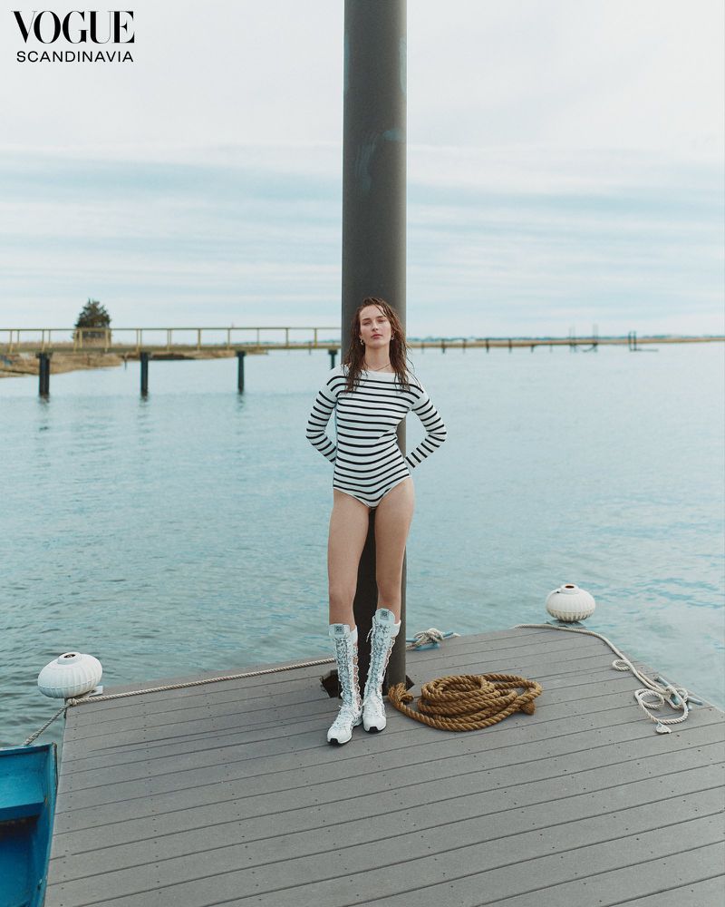 A young woman poses on a dock in a striped bodysuit and glitter boots, overlooking calm water beneath a cloudy sky, Vogue Scandinavia.