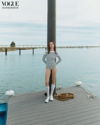 Model Julia Bergshoeff in a striped bodysuit and tall white boots standing on a wooden dock over water.