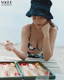 Model Julia Bergshoeff wearing a textured navy bucket hat and patterned bikini, leaning over a backgammon board on a wooden table.