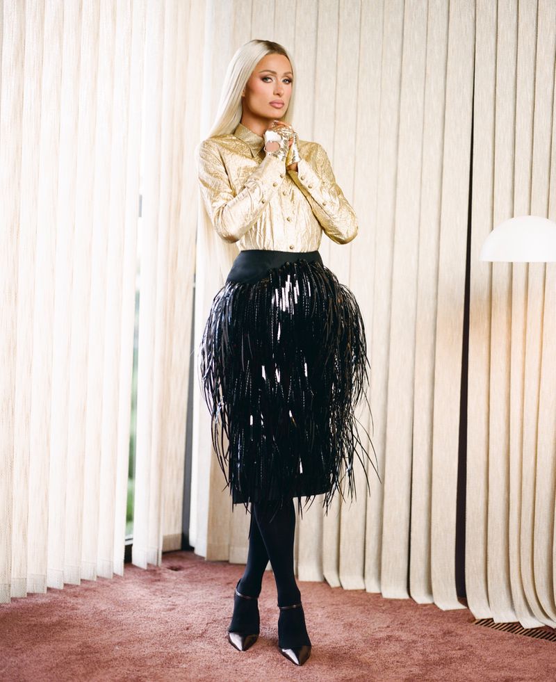 Paris Hilton in a gold blouse and black feathered skirt standing against vertical blinds for ODDA Magazine