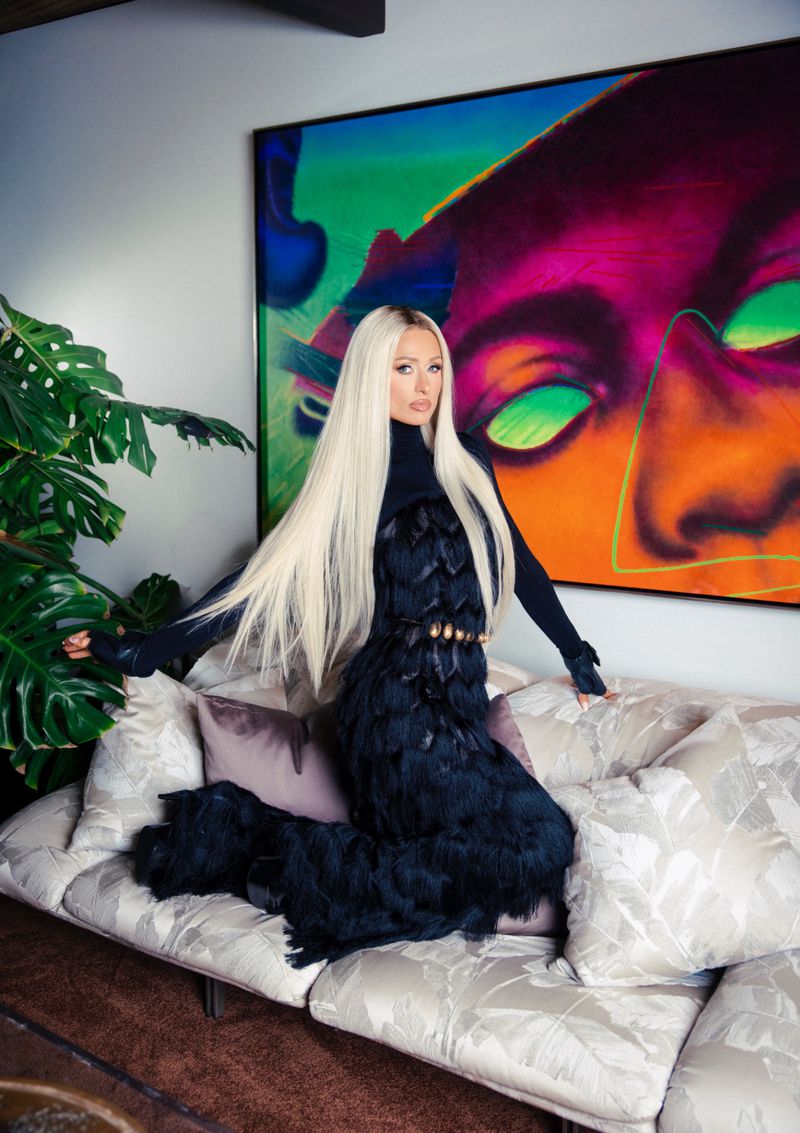 Paris Hilton with long blonde hair kneeling on a sofa wearing a black dress in an interior setting