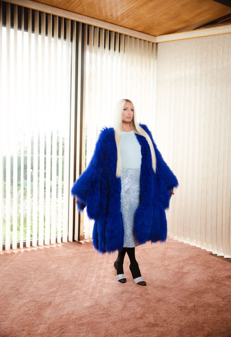 Paris Hilton wearing a vibrant blue fur coat and patterned skirt standing in a room with vertical blinds and a brown carpet