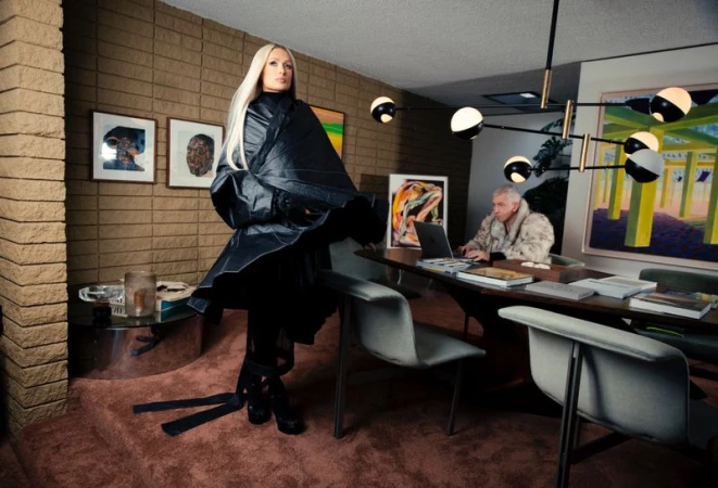 Paris Hilton wearing a black structured coat standing in a living room with an older man working at a dining table
