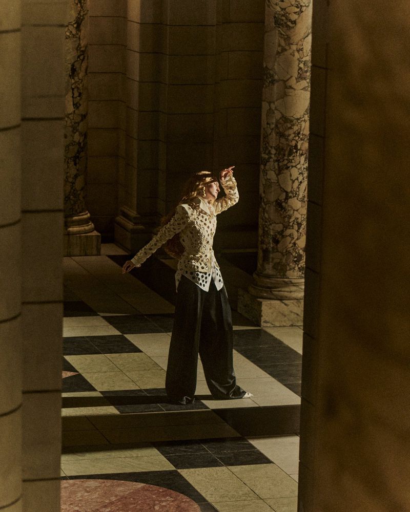 Model posing in ornate patterned top and dark trousers between neoclassical marble columns on a tiled floor.