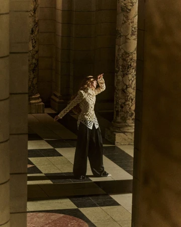 Model posing in ornate patterned top and dark trousers between neoclassical marble columns on a tiled floor.