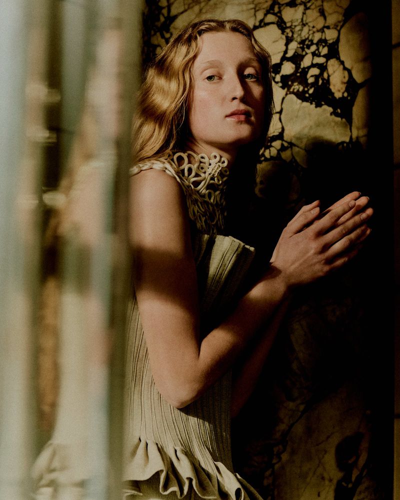 Model with long blonde wavy hair wearing a textured green dress with an ornate high collar against a marble background.