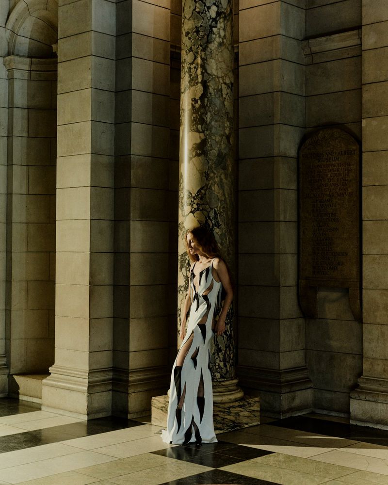 Model wearing a patterned white and black gown standing beside an ornate marble column inside a museum hall.