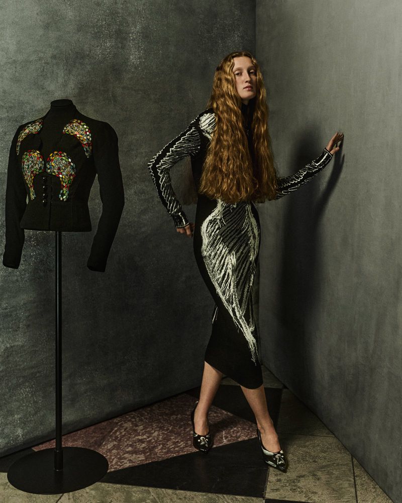 Model with long blonde hair in a black dress standing beside a mannequin on a stand against a dark textured wall.