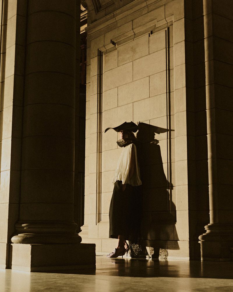 Model wearing dark wide-brimmed hat and pleated blouse posing against monumental stone column and block wall in warm low-key light