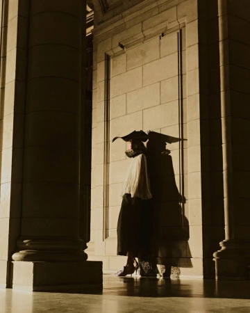 Model wearing dark wide-brimmed hat and pleated blouse posing against monumental stone column and block wall in warm low-key light