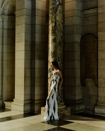 Model wearing a patterned white and black gown standing beside an ornate marble column inside a museum hall.