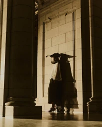 Model wearing dark wide-brimmed hat and pleated blouse posing against monumental stone column and block wall in warm low-key light
