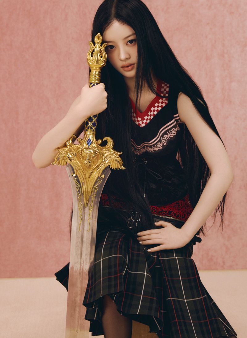 Young woman with long dark hair holding an ornate golden sword, wearing a patterned top and plaid skirt against a pink background.