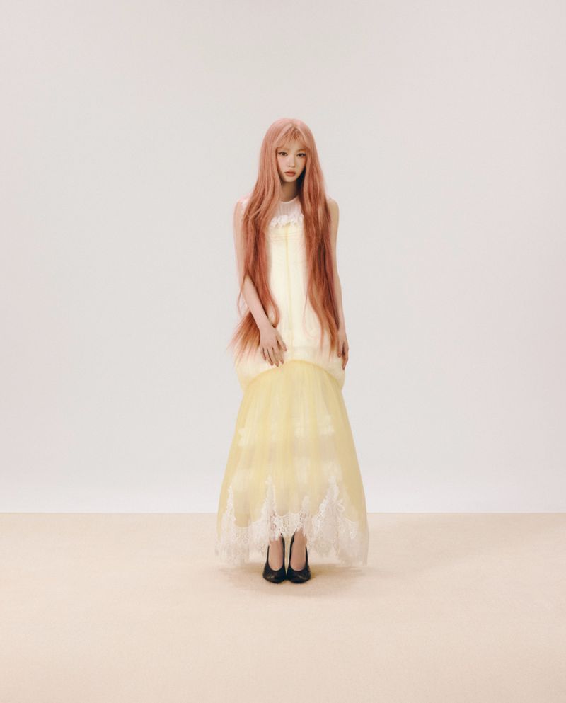 Full-length portrait of a woman with long pink hair wearing a layered yellow and white dress against a plain studio backdrop.