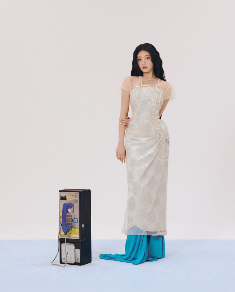Woman in an ornate white gown standing next to a vintage payphone against a neutral backdrop for ODDA Magazine.