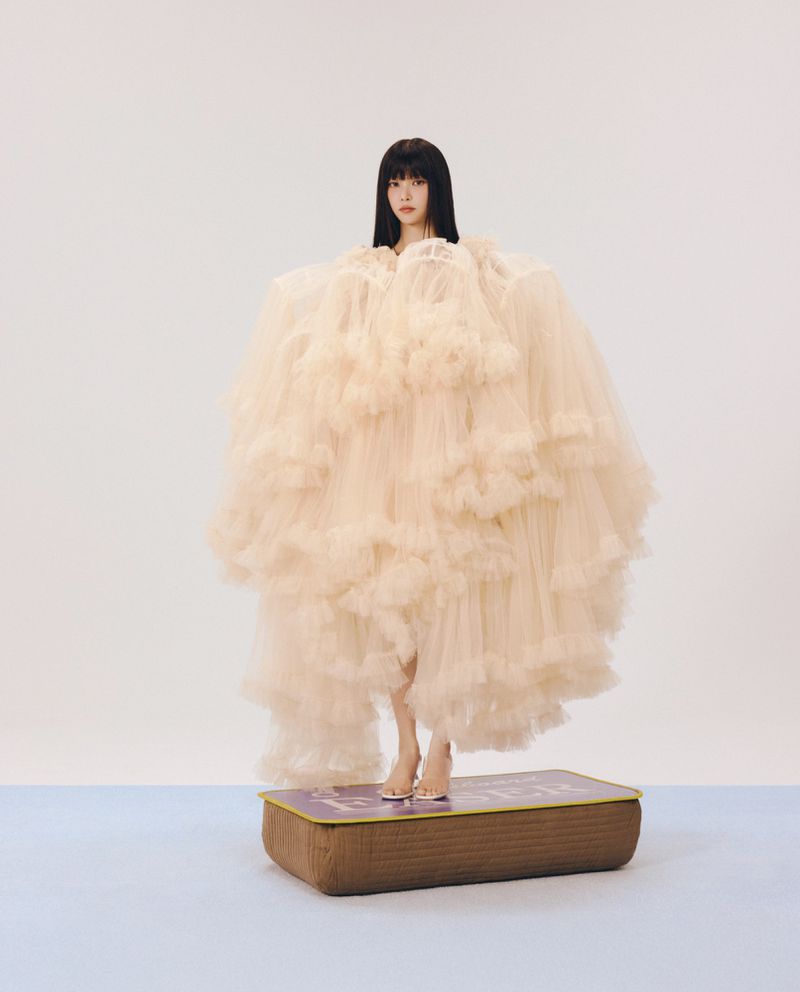A woman wearing a voluminous layered cream tulle dress standing on a book-shaped stool against a neutral backdrop