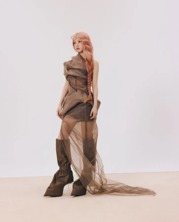 Woman with long braided pink hair wearing a sheer brown wrapped dress and tall brown boots against a light neutral background