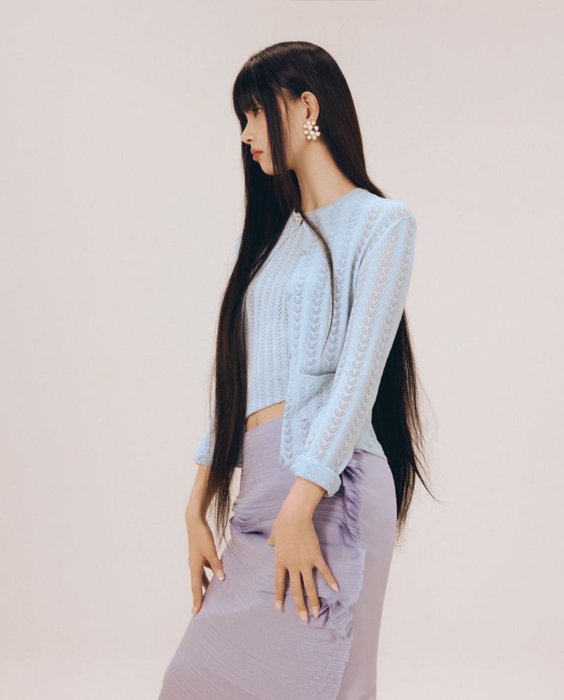 Profile view of woman with long dark hair wearing a light blue knit cardigan and purple skirt against a plain background