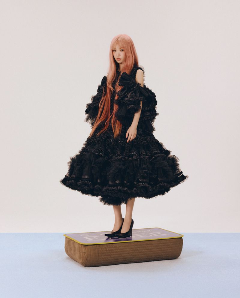 Woman with long pink hair wearing a voluminous black ruffled dress standing on a platform against a neutral background.