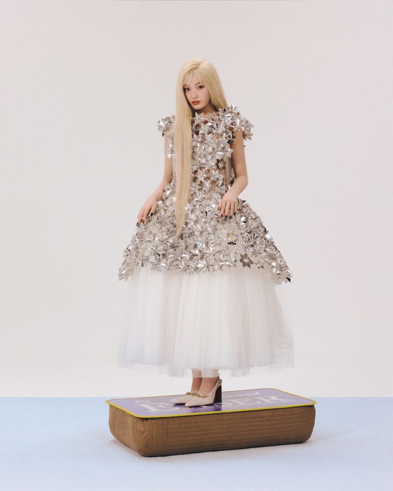Blonde woman in a silver metallic floral dress and white tulle skirt standing atop a large brown book-shaped prop against a white backdrop