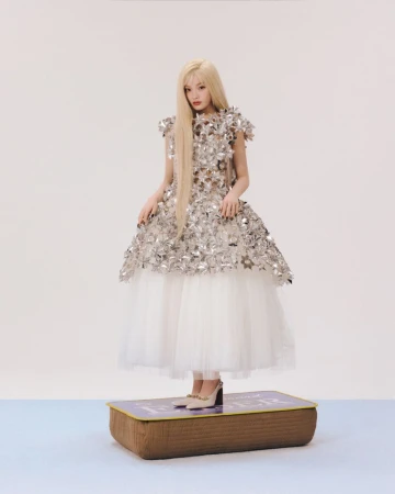 Blonde woman in a silver metallic floral dress and white tulle skirt standing atop a large brown book-shaped prop against a white backdrop