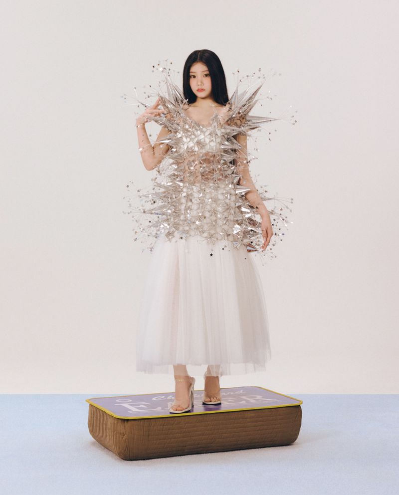 A woman in a metallic sculptural spiked top and white tulle skirt stands on a large matchbox prop.