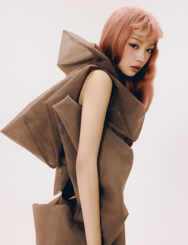 A woman with pink hair wearing a sculptural brown sheer garment against a plain white background for ODDA Magazine.