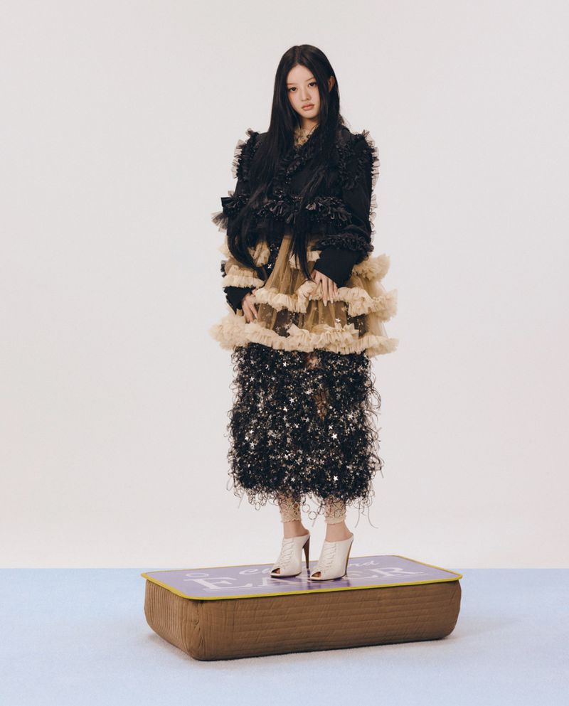 Young woman in a textured black ruffled dress standing on a low cushioned platform against a plain background