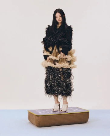 Young woman in a textured black ruffled dress standing on a low cushioned platform against a plain background