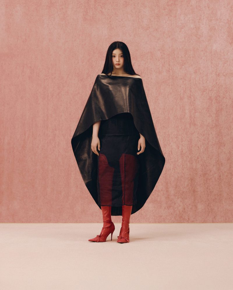 Woman with long dark hair wearing a sculptural black cape dress and red boots against a textured pink background