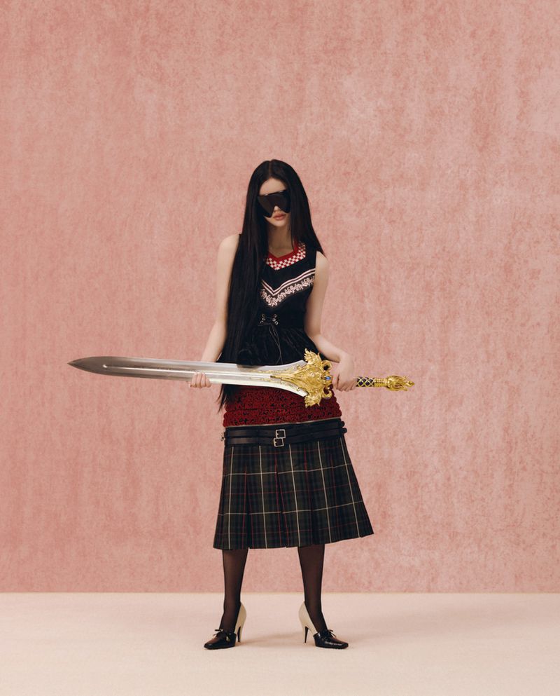 Model with long dark hair wearing a black dress and plaid skirt holding a large ornate sword against a pink background