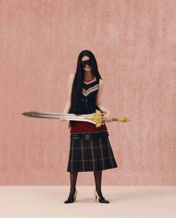 Model with long dark hair wearing a black dress and plaid skirt holding a large ornate sword against a pink background