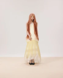 Full-length portrait of a woman with long pink hair wearing a layered yellow and white dress against a plain studio backdrop.