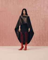 Woman with long dark hair wearing a sculptural black cape dress and red boots against a textured pink background