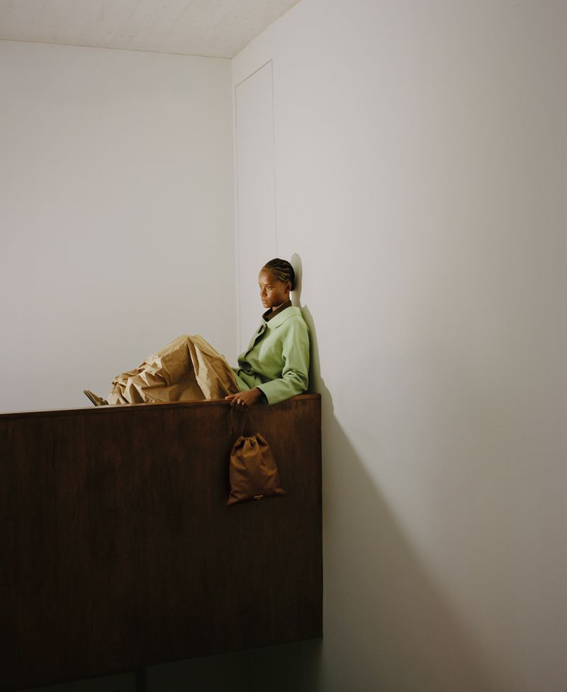 Letitia Wright seated against a white wall wearing a light green jacket and voluminous tan trousers for ODDA Magazine
