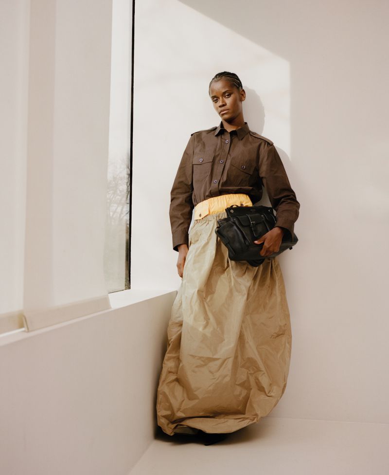 Letitia Wright wearing a brown button-up shirt and a voluminous tan skirt holding a black handbag against a wall