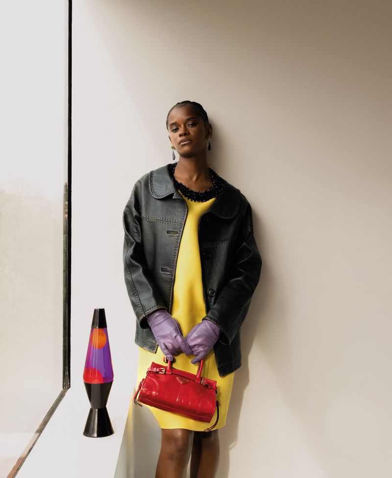 Letitia Wright wearing a yellow dress and black leather jacket holding a red handbag by a lava lamp for ODDA Magazine