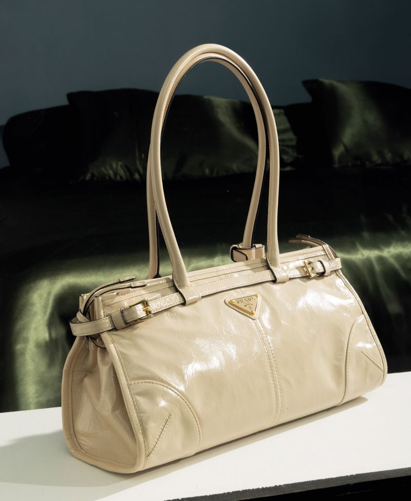 Beige patent leather handbag resting on a white surface with a dark green bed in the background.