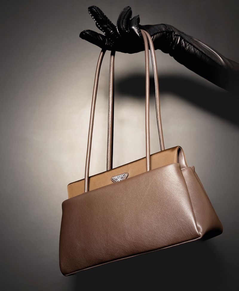 Gloved hand holding a brown leather shoulder bag against a neutral grey studio gradient background