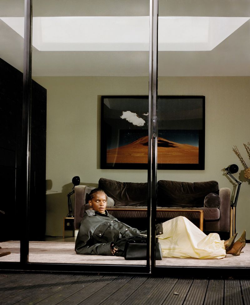 Letitia Wright reclining indoors in an olive green jacket and light skirt for ODDA Magazine, framed through glass doors.
