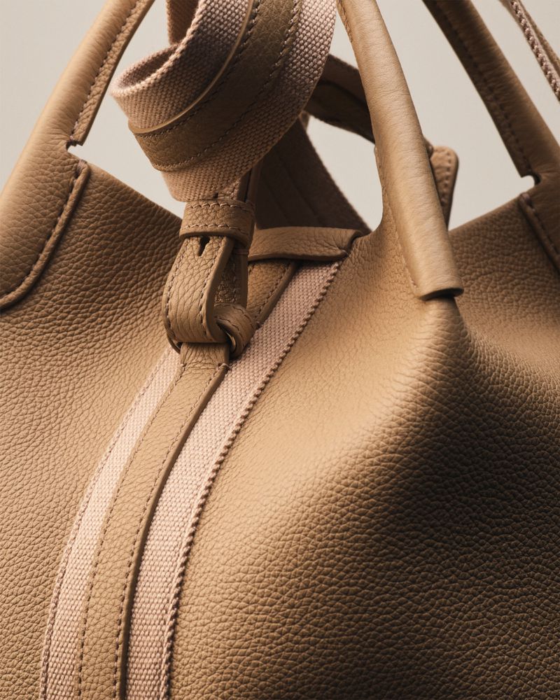 Close-up of a textured tan leather handbag with woven straps and buckle detail against a plain neutral background.